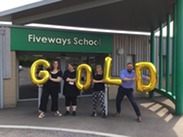 Teacher from Fiveways School standing outside the entrance holding golden lettered balloons that reads 'gold'.