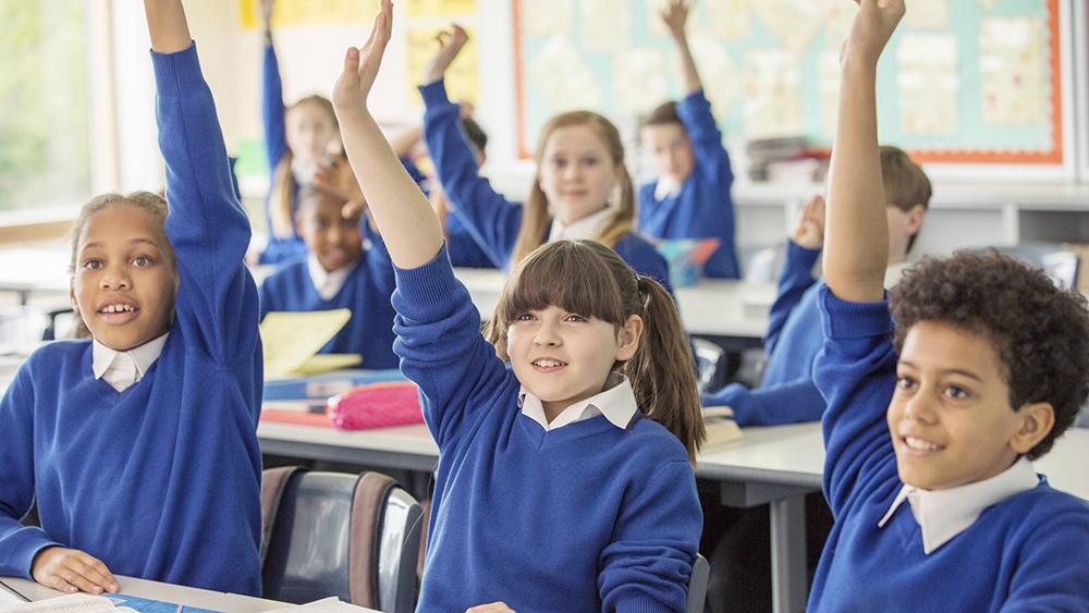 Elementary school children wearing blue school uniforms raising hands in classroom.