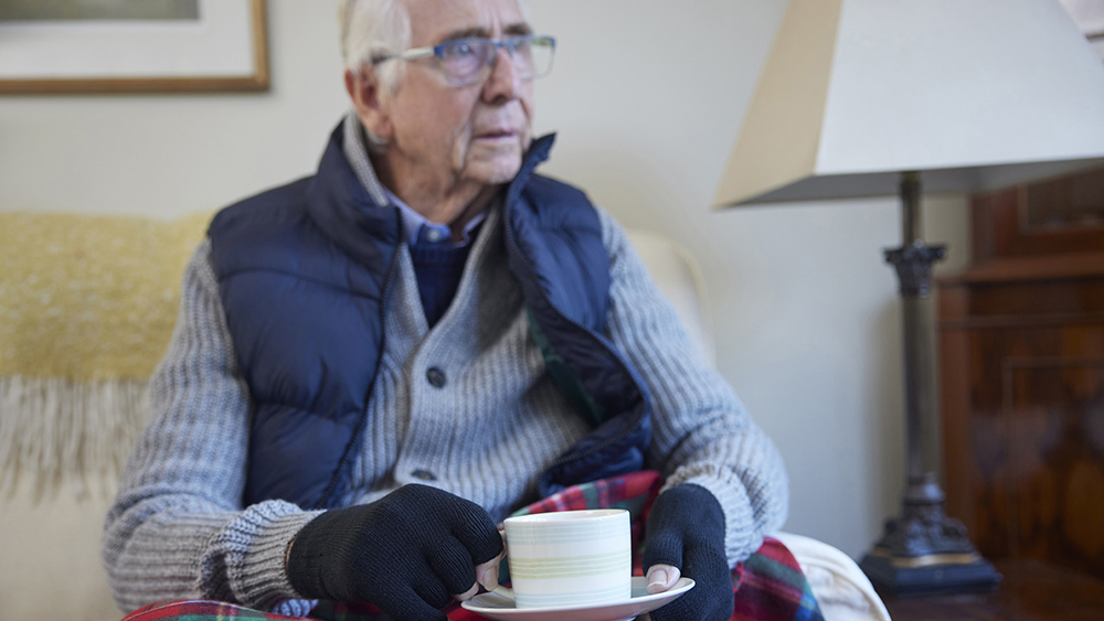 Senior man wearing extra clothes with hot drink trying to keep warm at home in energy crisis.