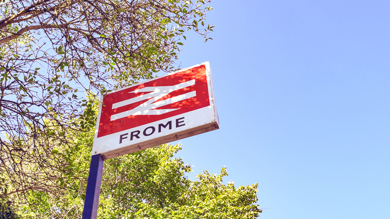 Low angle shot of a railway station sign for Frome with trees in the background.
