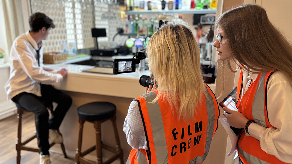 Female students directing a filming scene of a young man sat at a bar, who's probably too young to drink.