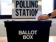 A hand posting a vote slip in black ballot box with a 'polling Station' sign in the background.