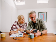 Man and woman sat at a table looking at a phone discussing finances, with paperwork, a calculator and a pair of glasses on the table.