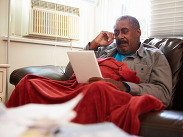 A black man wrapped in a blanket at home looking at his fuel bill.
