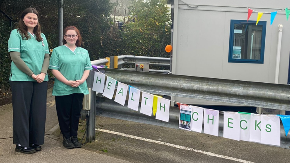 Two Somerset Council Public Health team members standing outside a business with bunting captioned: 'Health checks'.