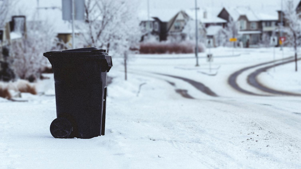 A black wheeled waste bin on the kerbside with snow and houses in the background by Michael Job Loquellano from Pexels.