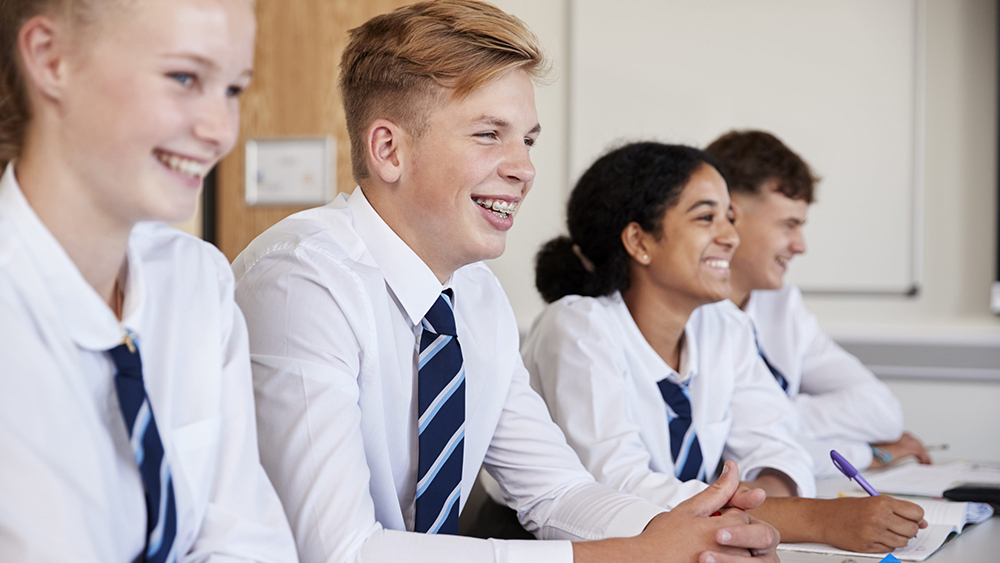 Line of secondary school students wearing uniforms sitting at their desks in a classroom.