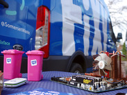 2 pink, Fixy-branded miniature wheelie bins with a mobile and PC motherboard on a table, with Fixy van in the background.