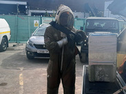 Beekeeper in protective suit stands by vehicle with equipment, near parked cars and green construction barriers.