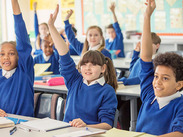 Students in blue uniforms raise their hands in a classroom, with notebooks and supplies on desks.