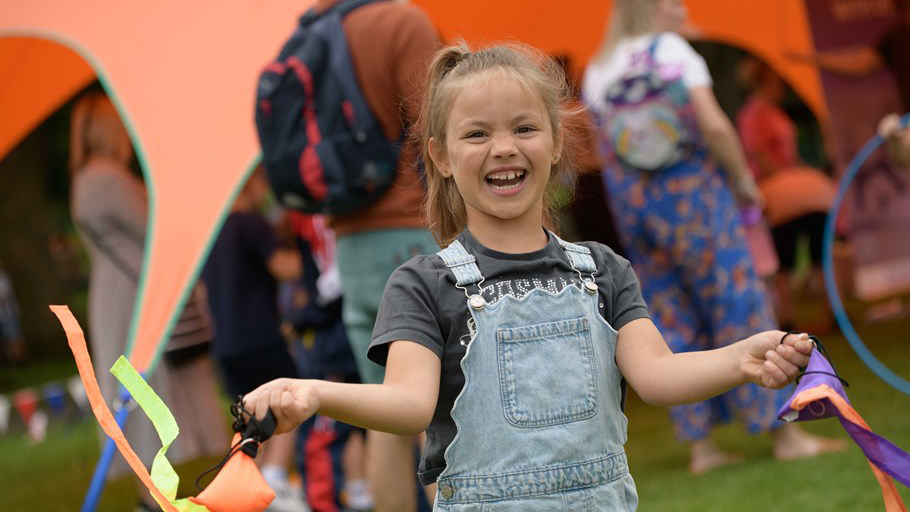 Child in denim overalls holding colourful ribbons at an outdoor event with people and a large orange tent in the background.