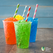 Four colourful slushies in plastic cups with striped straws on a wooden surface, with Food Standards Agency logo in the corner.