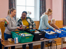 People organise and distribute food items at a food sharing event, with crates of supplies laid out on a table indoors.