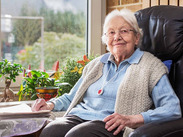 Elderly person sitting by a window wearing a Lifeline alarm, with potted plants nearby and greenery visible outside.