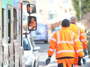 Waste collectors walking up a street with a recycling lorry following behind.
