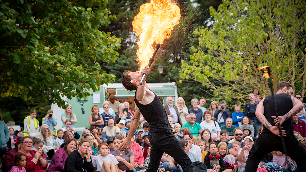 Fire performer breathes fire in front of a large seated audience at an outdoor event with trees and a green food truck in view.
