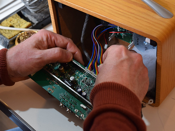 A mans hands opening the front of a radio to expose the PCB and wires with a soldering iron in the background.