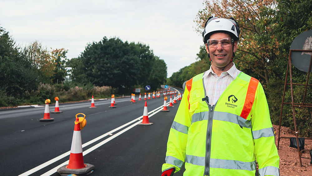 Cllr Richard Wilkins in a high-vis jacket and helmet on the A38 Chelston Link Road near Junction 26, with roadworks and cones behind.