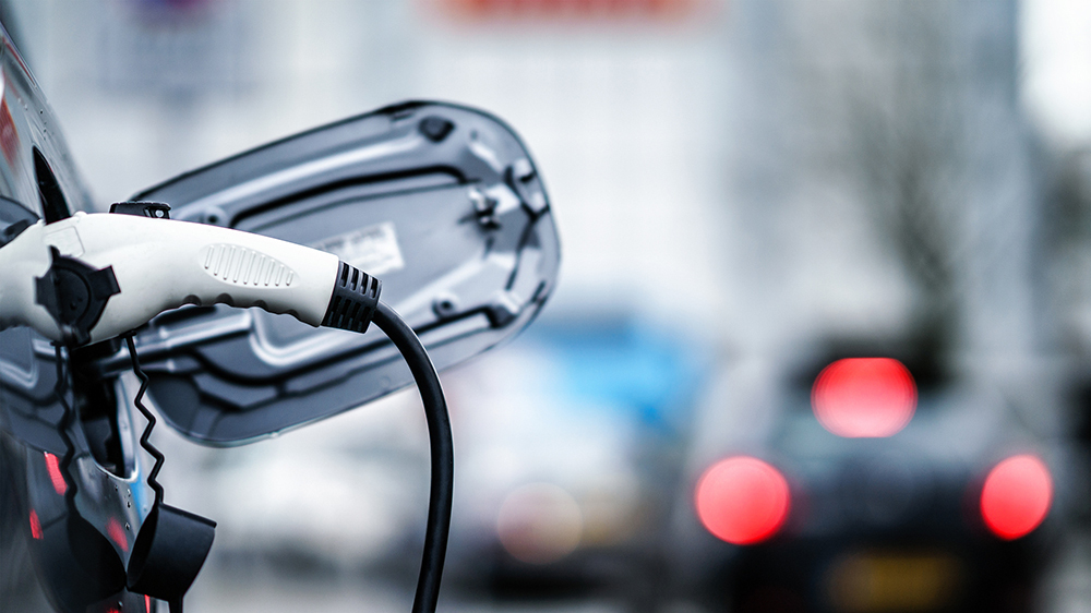 Close-up of an electric vehicle charging with a cable plugged into the port, blurred cars and lights in the background.
