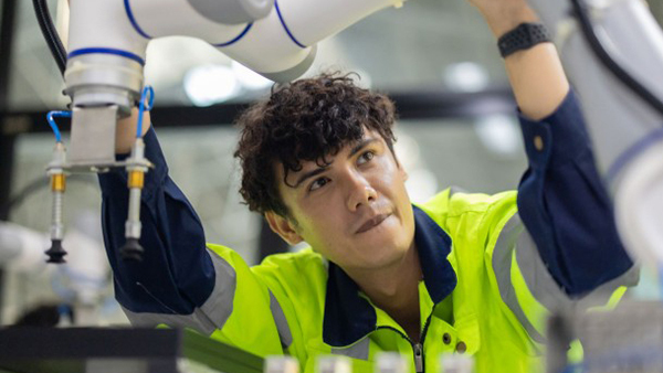 A young worker in a high-vis jacket operating a robotic arm in an industrial setting.