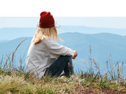 A woman in a red beanie and white sweater sits cross-legged on a grassy hill, gazing at distant blue mountains.