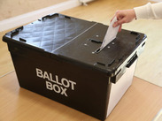 "A hand placing a ballot into a black ballot box labelled 'BALLOT BOX' in white letters."
