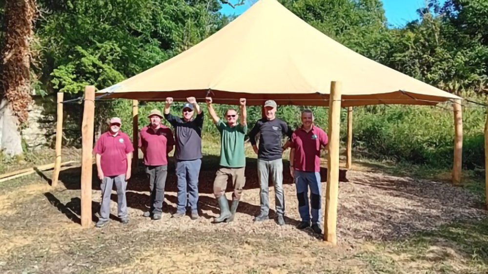 Ham Hill activity hub with six people gathered under a large beige canopy in a wooded outdoor setting.