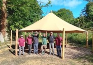 Ham Hill activity hub with six people gathered under a large beige canopy in a wooded outdoor setting.
