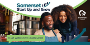Two people smiling at a laptop in a cafe with 'Somerset Start Up and Grow' and 'Chat with us to learn more' text.