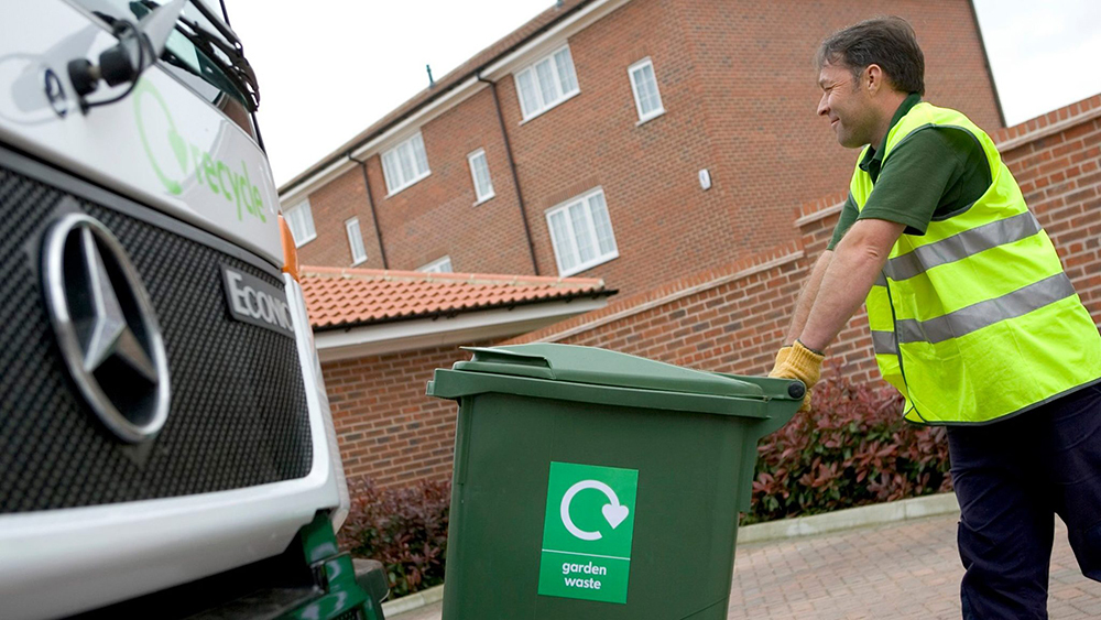 A worker in a reflective vest pushes a green garden waste bin towards a recycling truck in front of residential buildings.