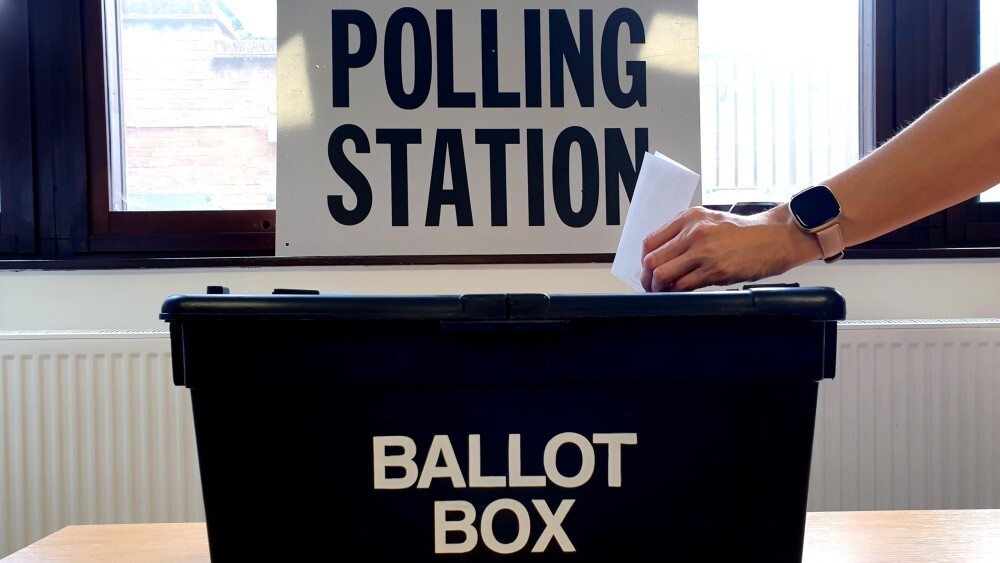 A hand posting a vote slip in black ballot box with a 'polling Station' sign in the background.