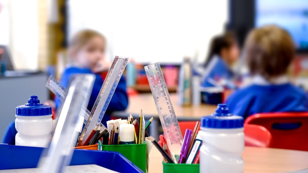 Classroom scene with focus on rulers, pencils, and water bottles on a table; blurred background shows children in blue uniforms.