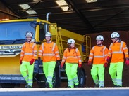 Somerset Council and Kier workers in high-vis gear walk past a yellow gritter truck inside a winter service depot.