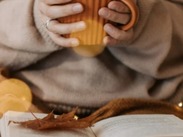 Person holding an orange mug with both hands, wearing a cosy sweater, with an open book and dried leaf in the foreground.