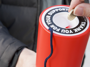 Hand placing a coin into a red charity collection tin with “Thank you for your support” on the lid.