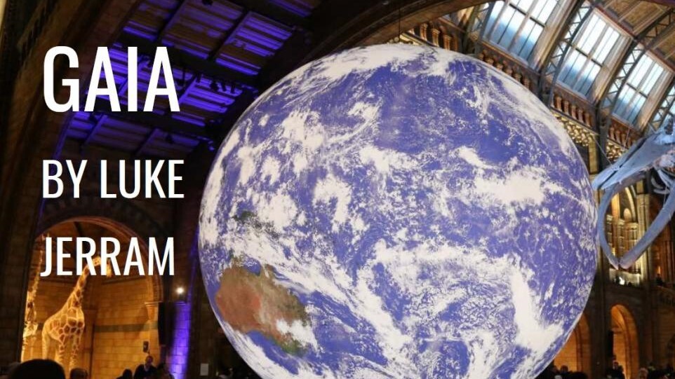 Large illuminated Earth sculpture titled “Gaia” by Luke Jerram displayed inside a grand hall with arched windows.