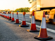 Row of orange traffic cones along a road with vehicles passing, indicating roadworks or lane closure at sunset.