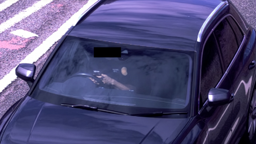 Front view of a dark Audi car on a road, driver visible through windscreen holding a mobile phone while driving.