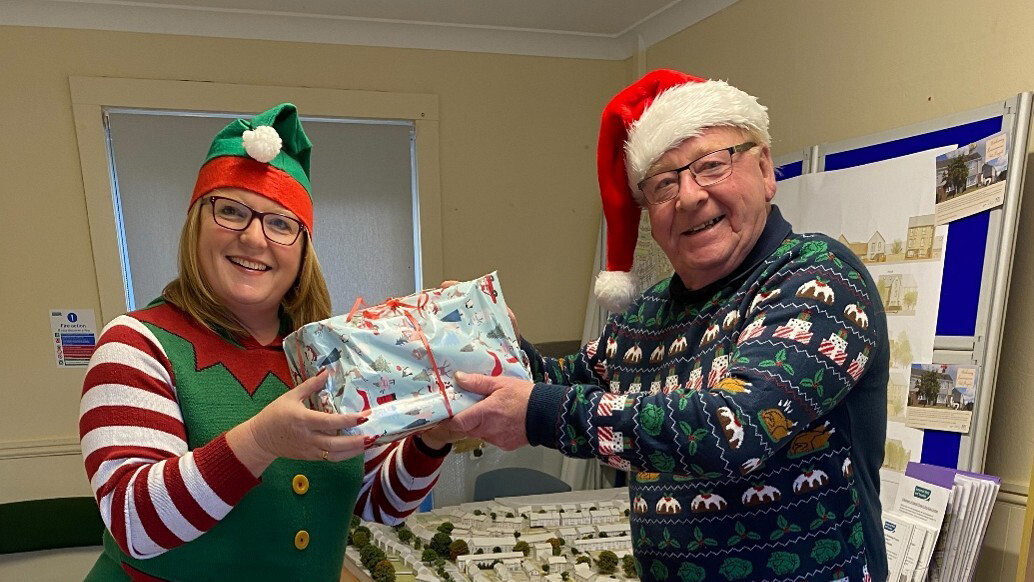 2 people in festive attire exchange a wrapped Christmas gift, supporting the Christmas Sparkle Project for children in need.