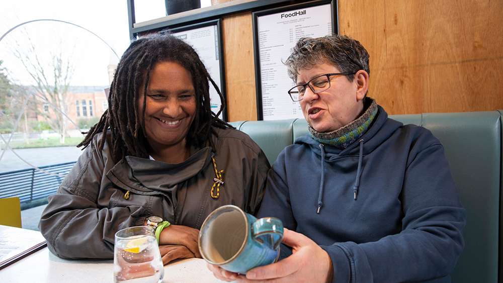 Two people sit at a table laughing together. One is holding a mug and the other is looking at it.