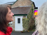 Two people using a thermal imaging camera on a smartphone to check heat loss from a stone house with tiled roof and white door.