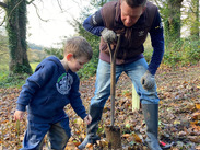 Adult and child planting a tree with a spade among fallen leaves during Ham Hill’s National Tree Week habitat-connecting project.