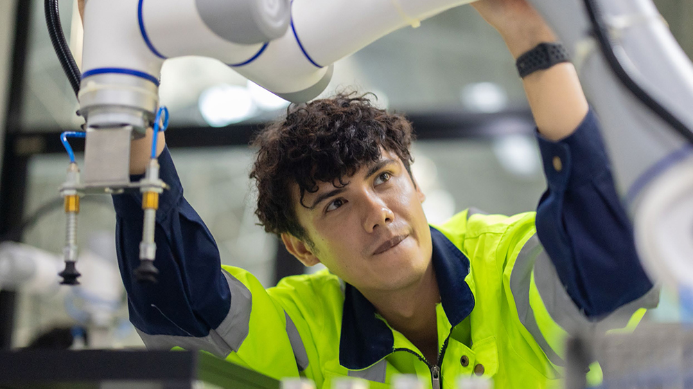 A young worker in a high-vis jacket operating a robotic arm in an industrial setting.