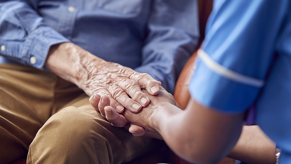 Close up of a female care worker in uniform holding the hands of a senior man sitting in a care home lounge.