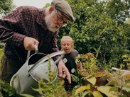 An older person watering garden plants with a child beside them in a lush, green outdoor setting.