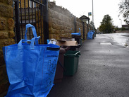 Recycling bags, boxes and bins lined up along a wet pavement beside a stone wall on a residential street.
