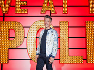 Comedian Stuart Goldsmith Person standing in front of the bright red Live at the Apollo stage backdrop with large illuminated lettering.