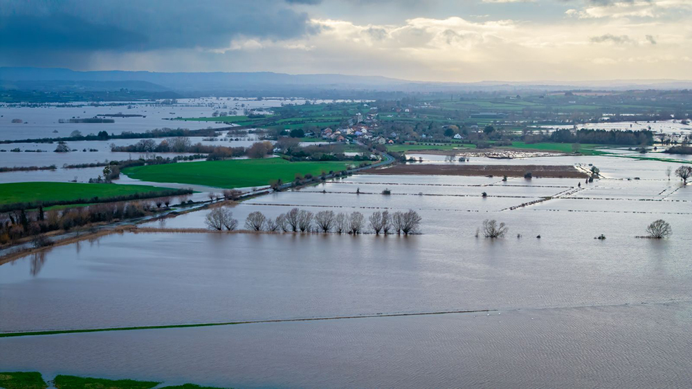 Aerial view of flooding across fields in the Moorland area, with waterlogged farmland, scattered trees and distant villages under cloudy skies.