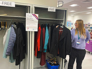 Free winter coat exchange in a library, with coats hanging on open racks and a staff member standing beside them.