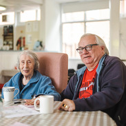 2 older adults sit at a table with mugs in a bright communal lounge, chatting and relaxing, from Age Positive Image Library.